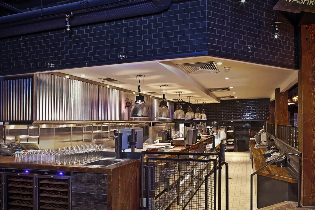 Modern industrial bar and kitchen pass with warm lighting and exposed ductwork.