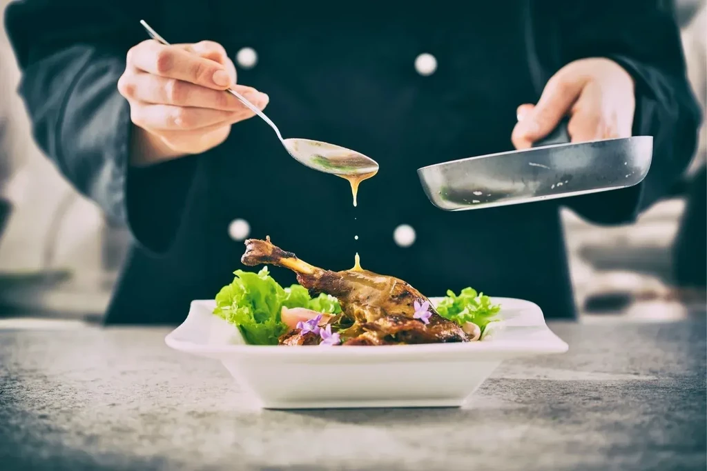 Chef's hands drizzle sauce on roasted meat with lettuce and purple flowers.