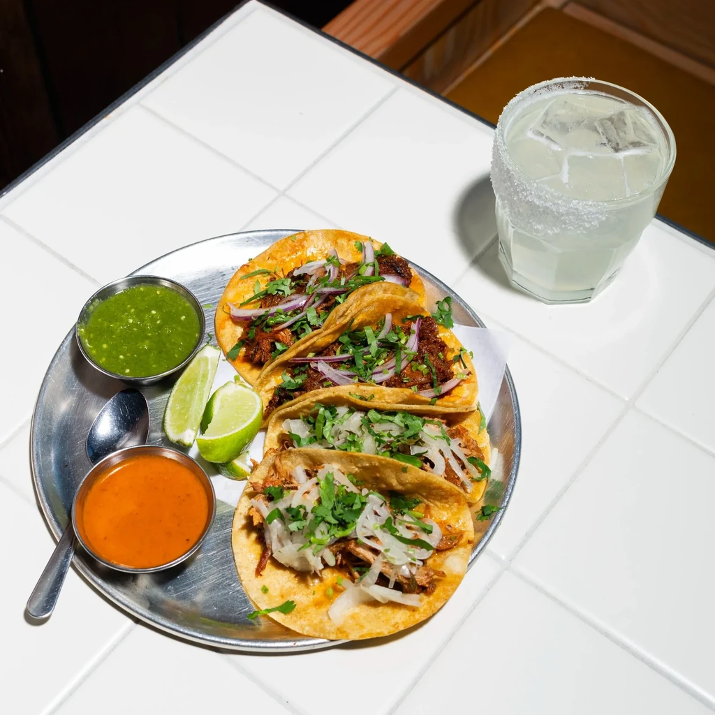 A metal plate of four tacos, two salsas, lime, and a cocktail.