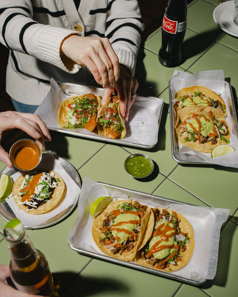 Top-down view of hands with tacos and drinks on a tiled table.
