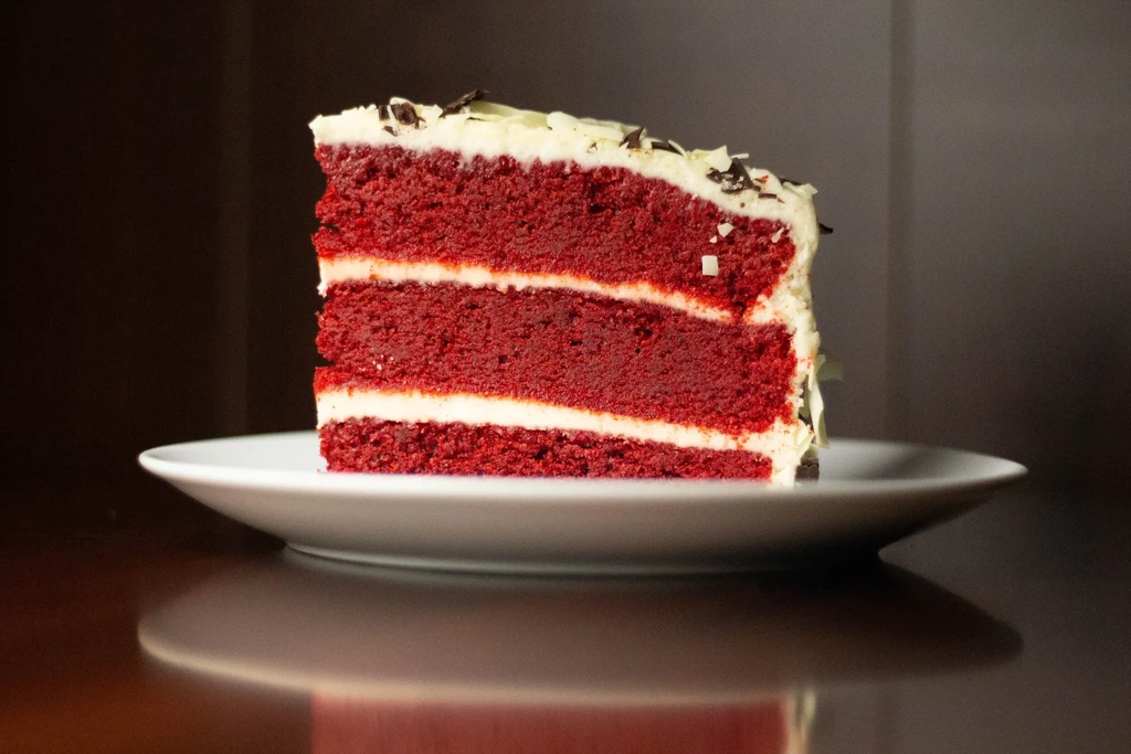 Red velvet cake slice with white frosting and chocolate shavings on plate.