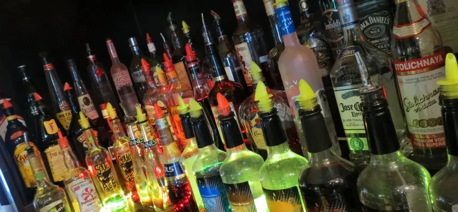 Tightly packed liquor bottles with colorful pour spouts on a backlit bar shelf.