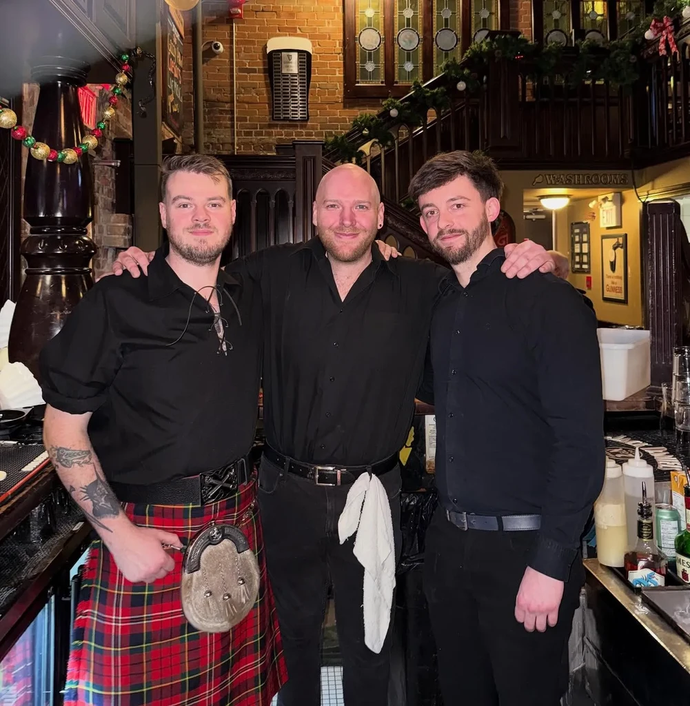 Three smiling pub staff, one in a kilt, in a bar.