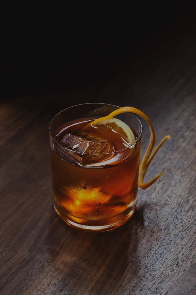 Amber cocktail in rocks glass, garnished with orange peel and large ice.