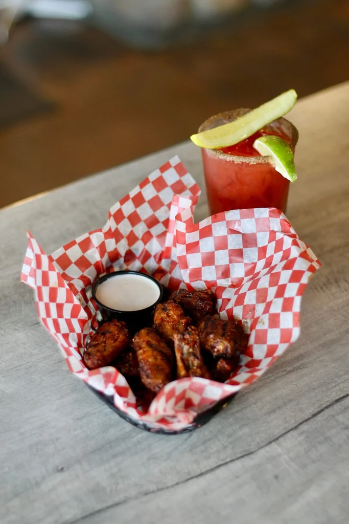 Chicken wings in basket, white dip, and garnished cocktail on wood.