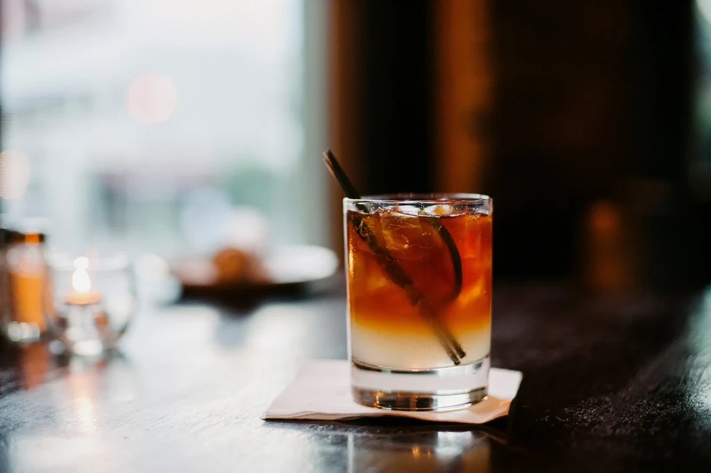 Layered cocktail with ice, green garnish, and straw on dark wooden table.