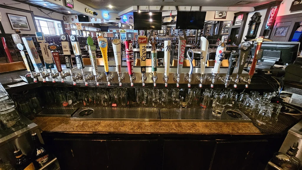 Long line of 25 diverse beer taps above empty glasses at a bar.