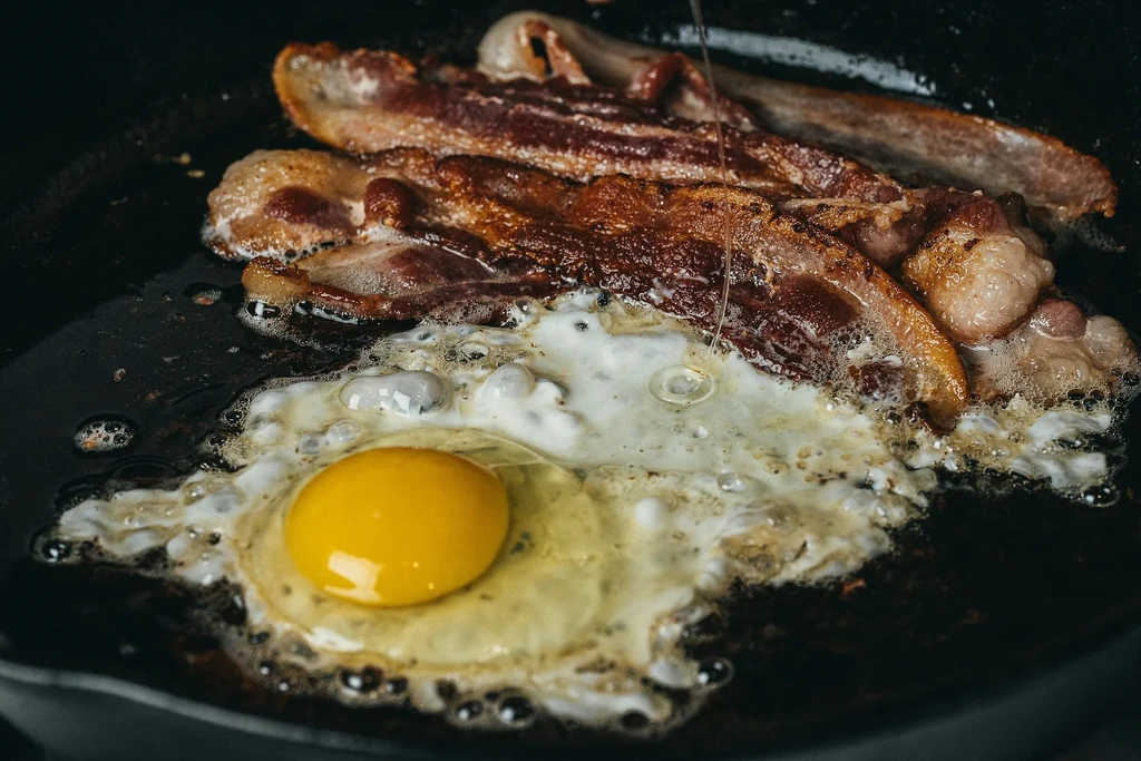 Well-rendered bacon and a sunny-side-up egg frying in a dark pan.