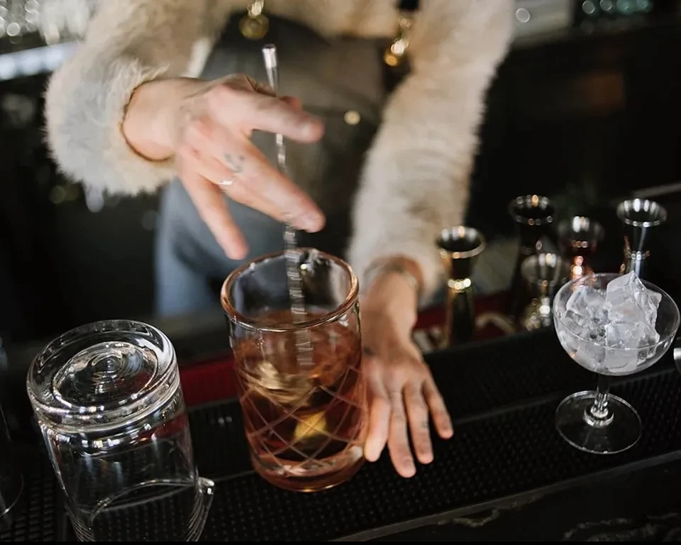 Bartender's fluffy-sleeved hands stir an amber cocktail with ice.