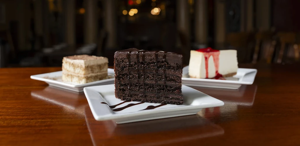 Three desserts: focused chocolate cake, tiramisu, and cheesecake on a wooden surface.