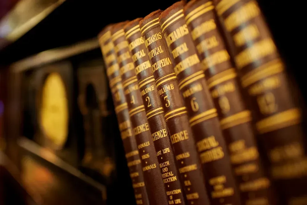 Close-up of vintage, leather-bound books with gold lettering on dark shelf.