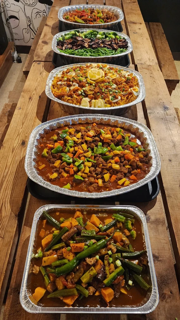 Six foil trays of various prepared dishes on a wooden table.