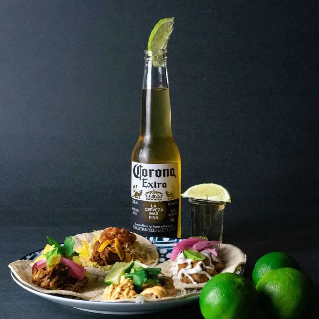 Five colorful tacos, Corona beer with lime, shot glass, and limes.