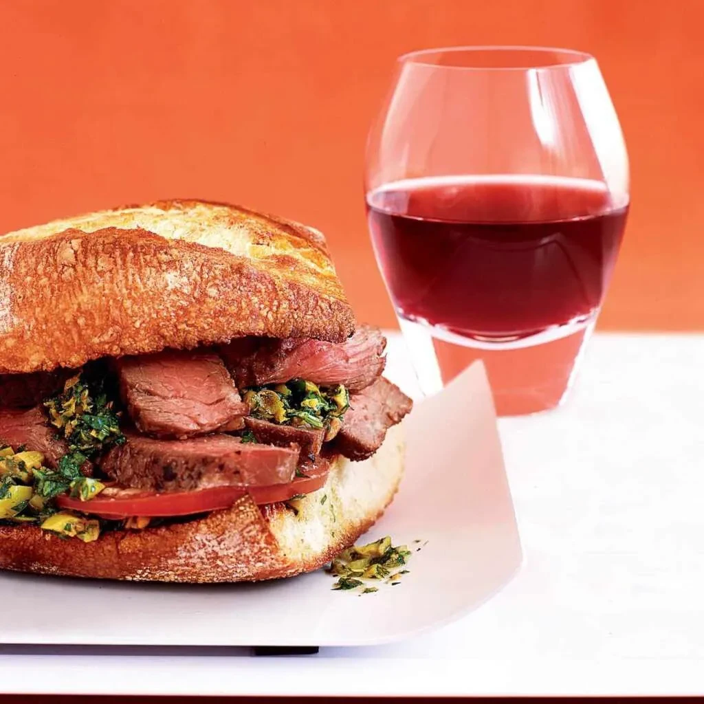 Rustic sandwich with rare beef, chimichurri, tomato, and red beverage.