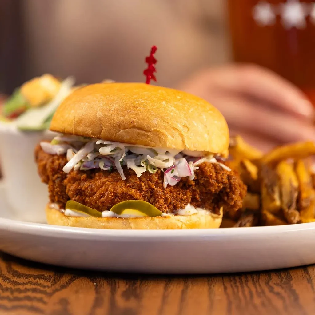 Fried chicken sandwich, coleslaw, pickles, and waffle fries on a wooden table.