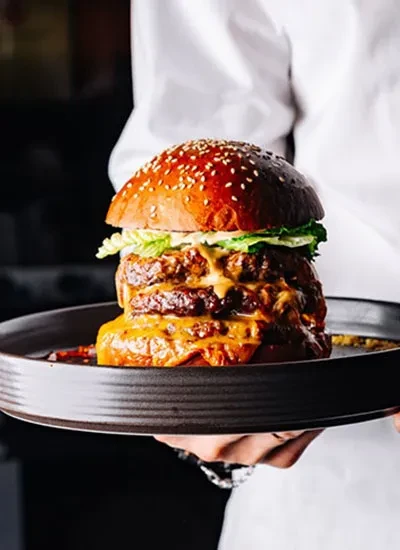 Large multi-patty burger with melted cheese, lettuce, held on dark plate.