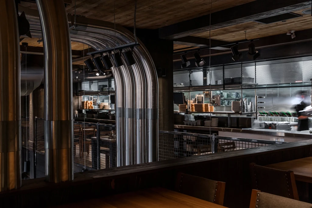 Modern industrial restaurant with prominent curved ducts and bright open kitchen.