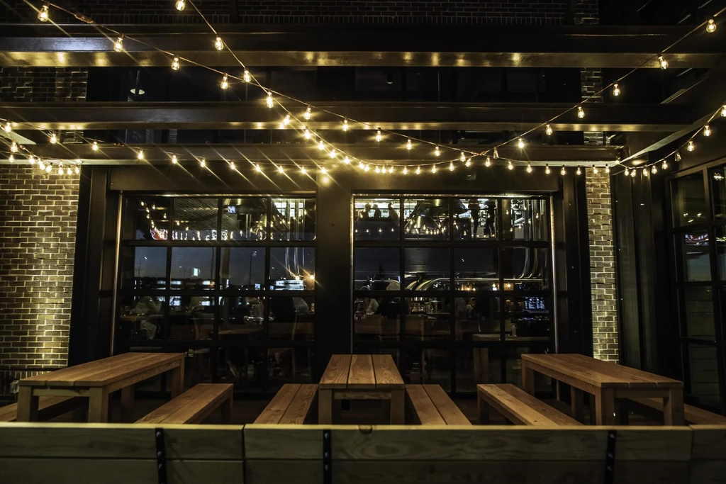 Nighttime outdoor patio with wooden picnic tables and glowing bistro lights.