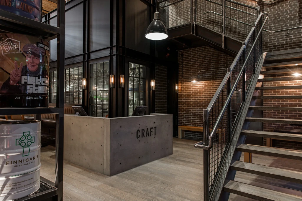 Industrial bar interior, beer kegs, 'CRAFT' counter, brick walls, and staircase.