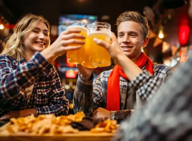 Smiling man and woman in plaid shirts toast large beers at a bar.