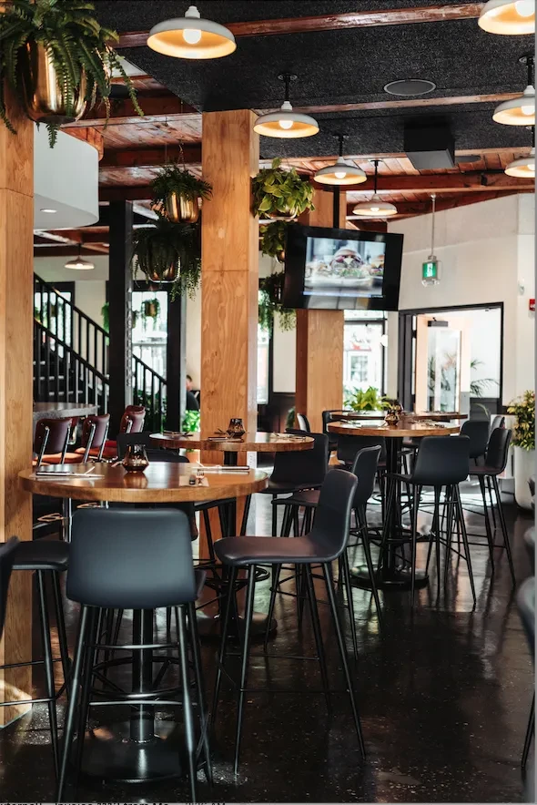 Warmly lit modern restaurant with wooden beams, high-top tables, and hanging plants.