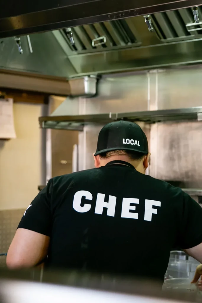 Chef's back in kitchen, 'CHEF' shirt, 'LOCAL' cap, stainless steel background.