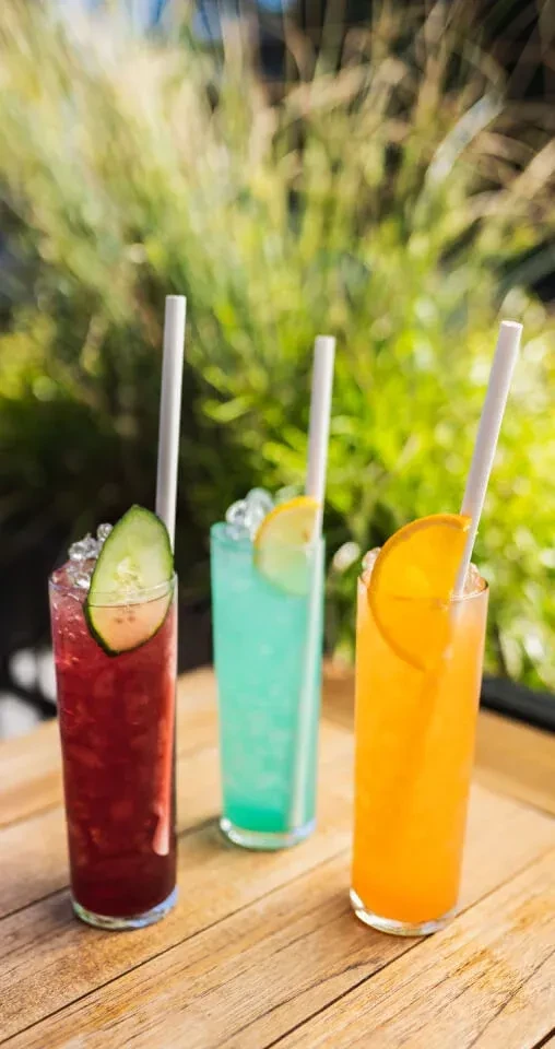 Three colorful drinks with ice, fruit, and straws on wood.
