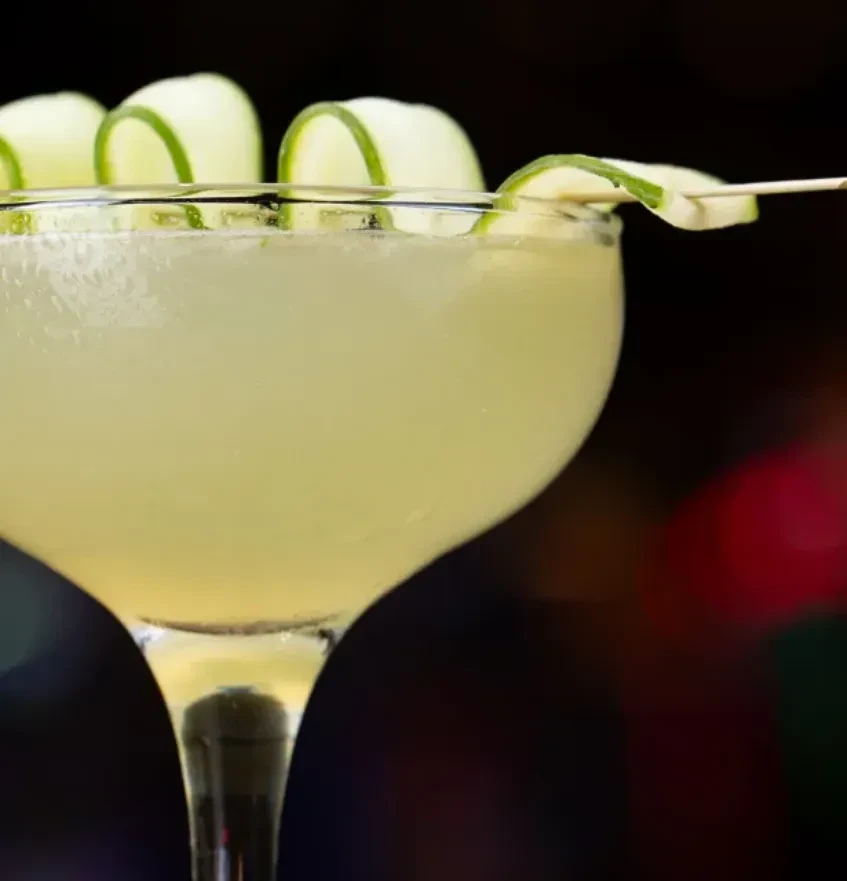 Pale green cocktail in coupe glass, garnished with curled cucumber.