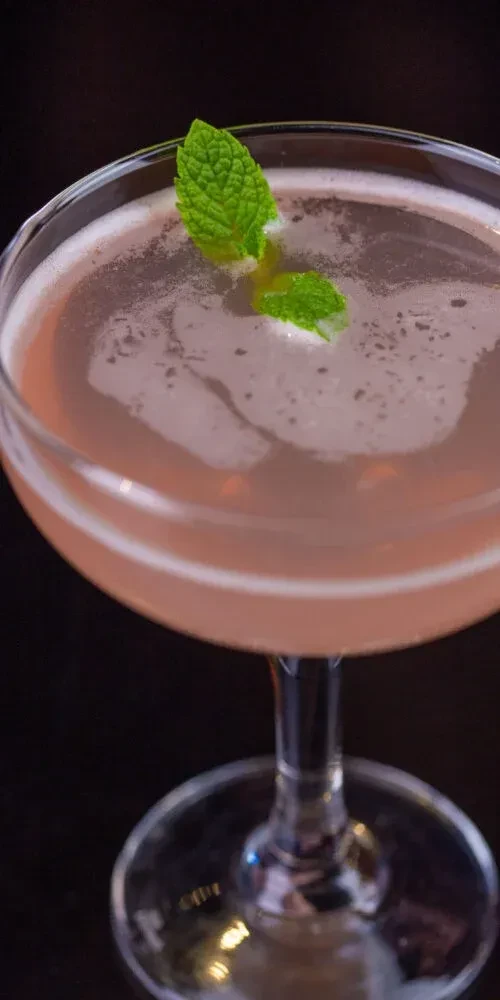 Pink frothy cocktail in a stemmed glass, garnished with mint leaves.