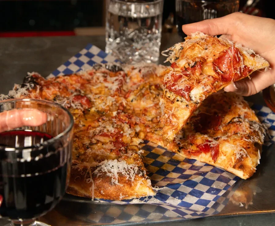 Hand lifts pizza slice with generous toppings, cheese, and red wine.