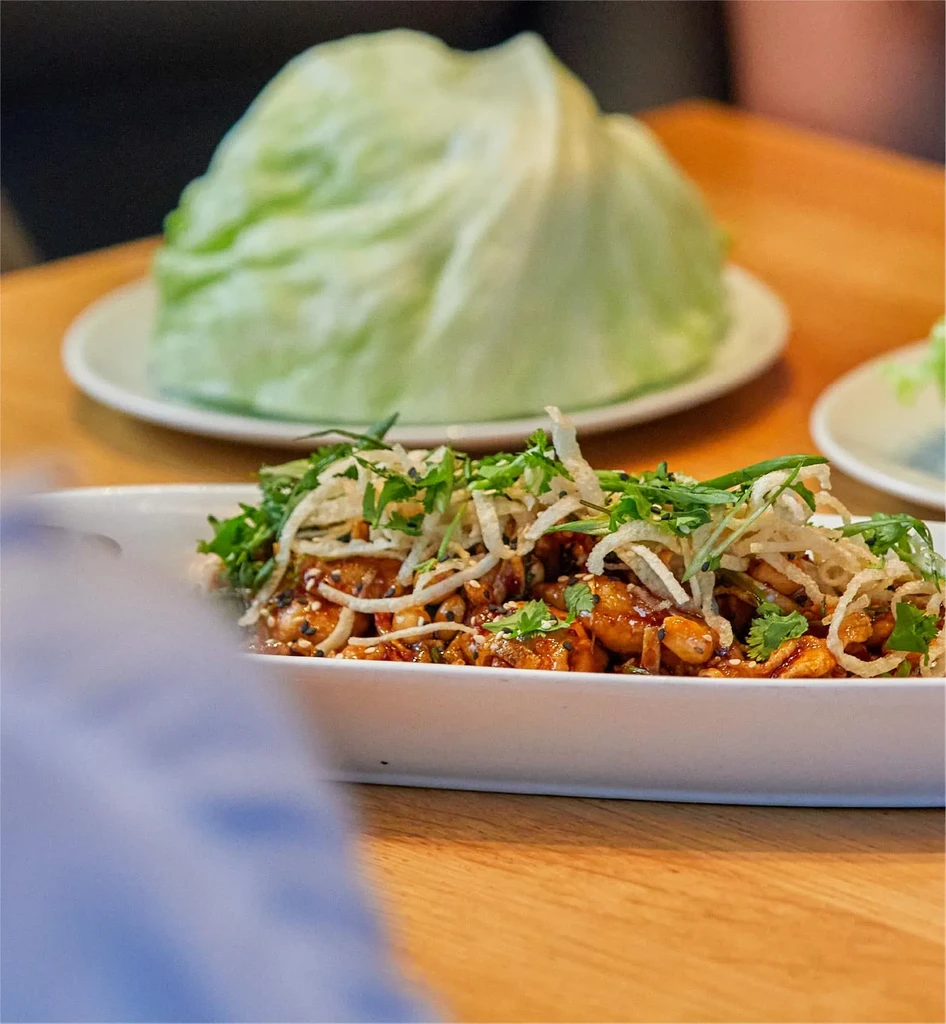 Close-up of savory protein, nuts, herbs, and lettuce wraps on wood.