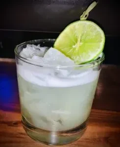 Close-up of a pale green cocktail with crushed ice, foam, and lime.