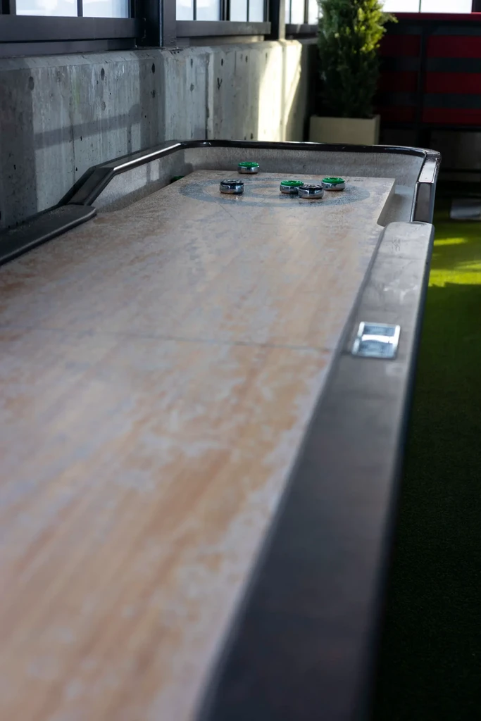 Light wood shuffleboard table with pucks, concrete wall and windows.