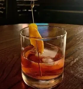Amber-reddish cocktail in rocks glass, ice, orange peel, wood.