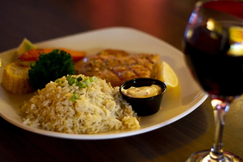 A grilled protein meal with rice, vegetables, sauce, and red wine.