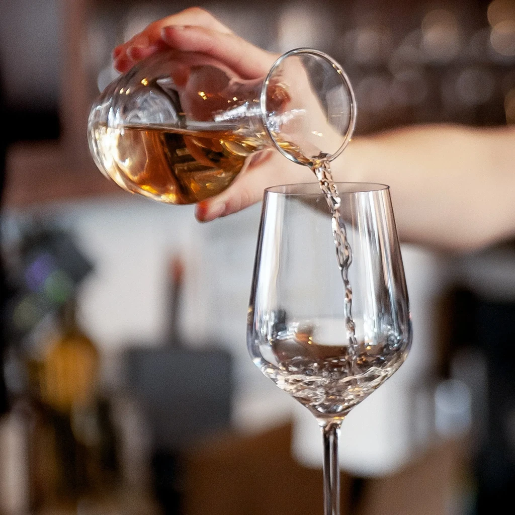 Close-up of hand pouring amber liquid from carafe into wine glass.