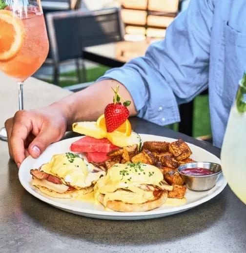 Brunch plate with Eggs Benedict, potatoes, fruit, and drinks on a patio.