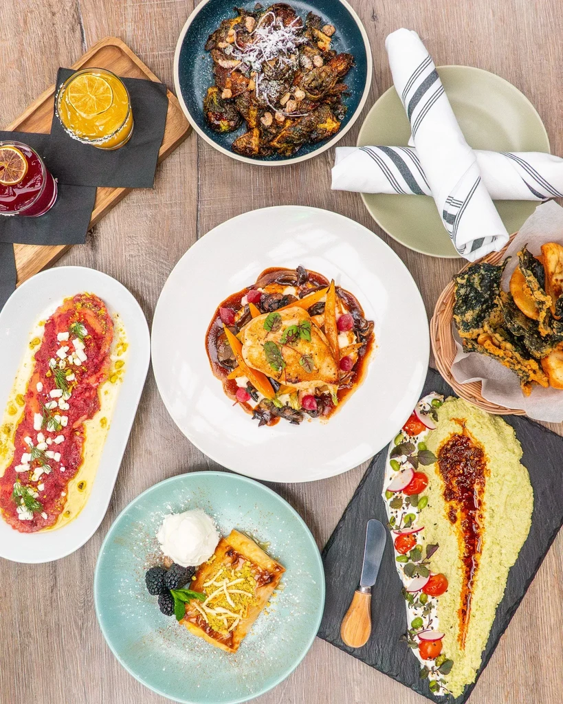 Top-down view of diverse restaurant dishes and cocktails on a wooden table.