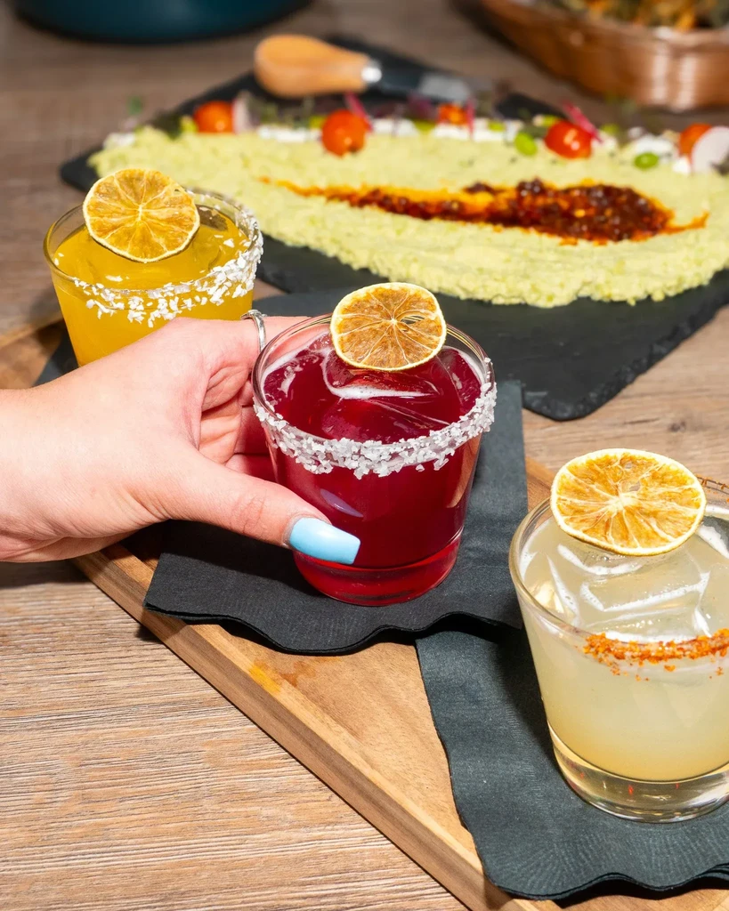 Three colorful, garnished drinks on a wooden board with a green dip.