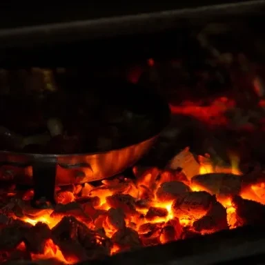 Metal pan cooking food over glowing red-orange hot coals.