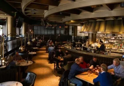 Industrial-chic restaurant interior with a bar, warm lighting, and many patrons.