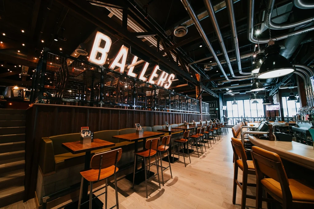 Industrial-style restaurant interior with "BALLERS" sign, olive-green booths, and bar.
