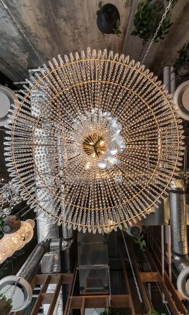 Circular crystal chandelier against distressed industrial ceiling with plants.