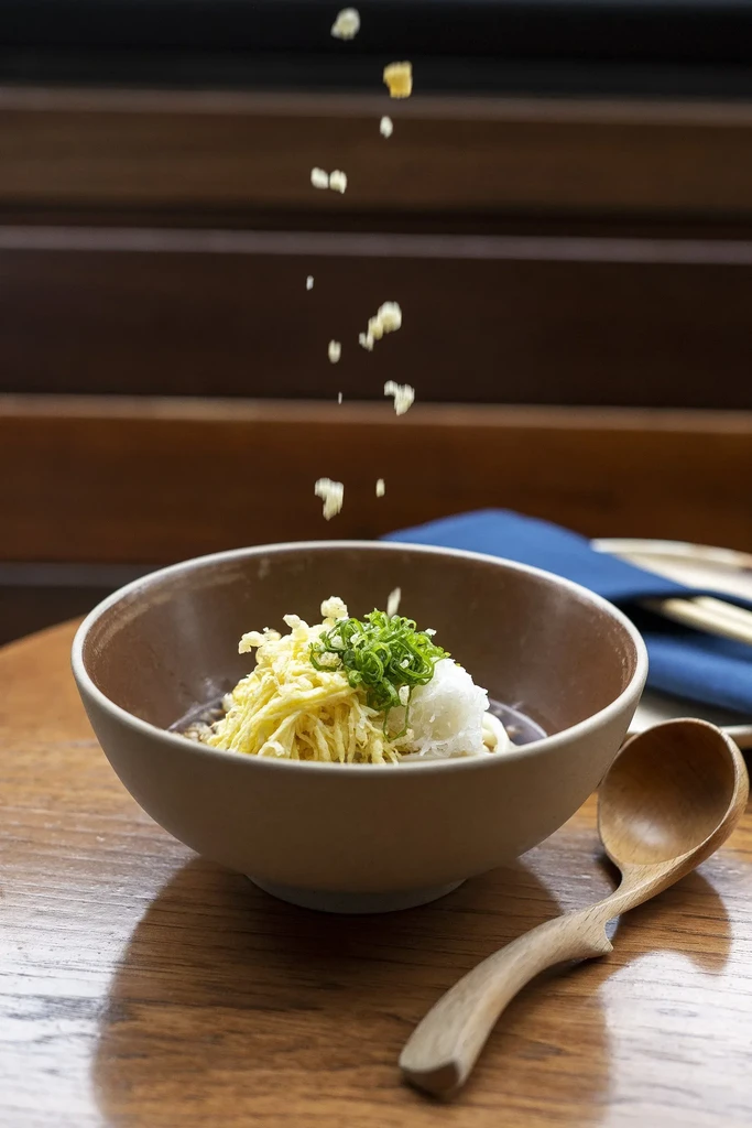 Close-up of Japanese noodles with toppings and falling flakes on wooden table.