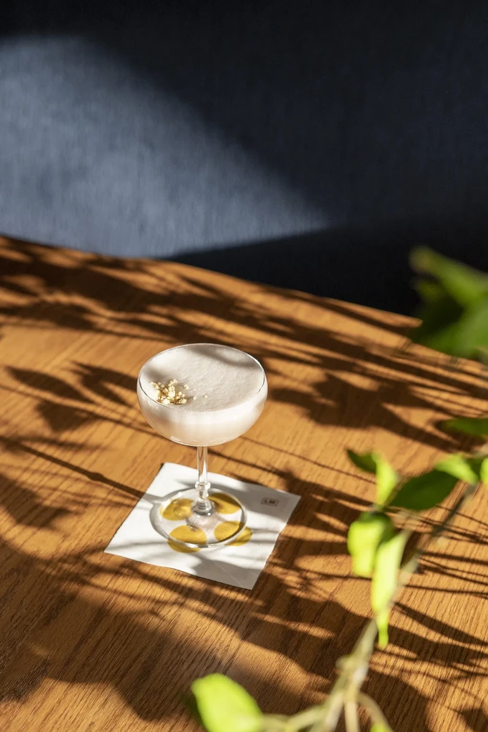 Off-white cocktail in coupe glass on a shadowed wooden table.