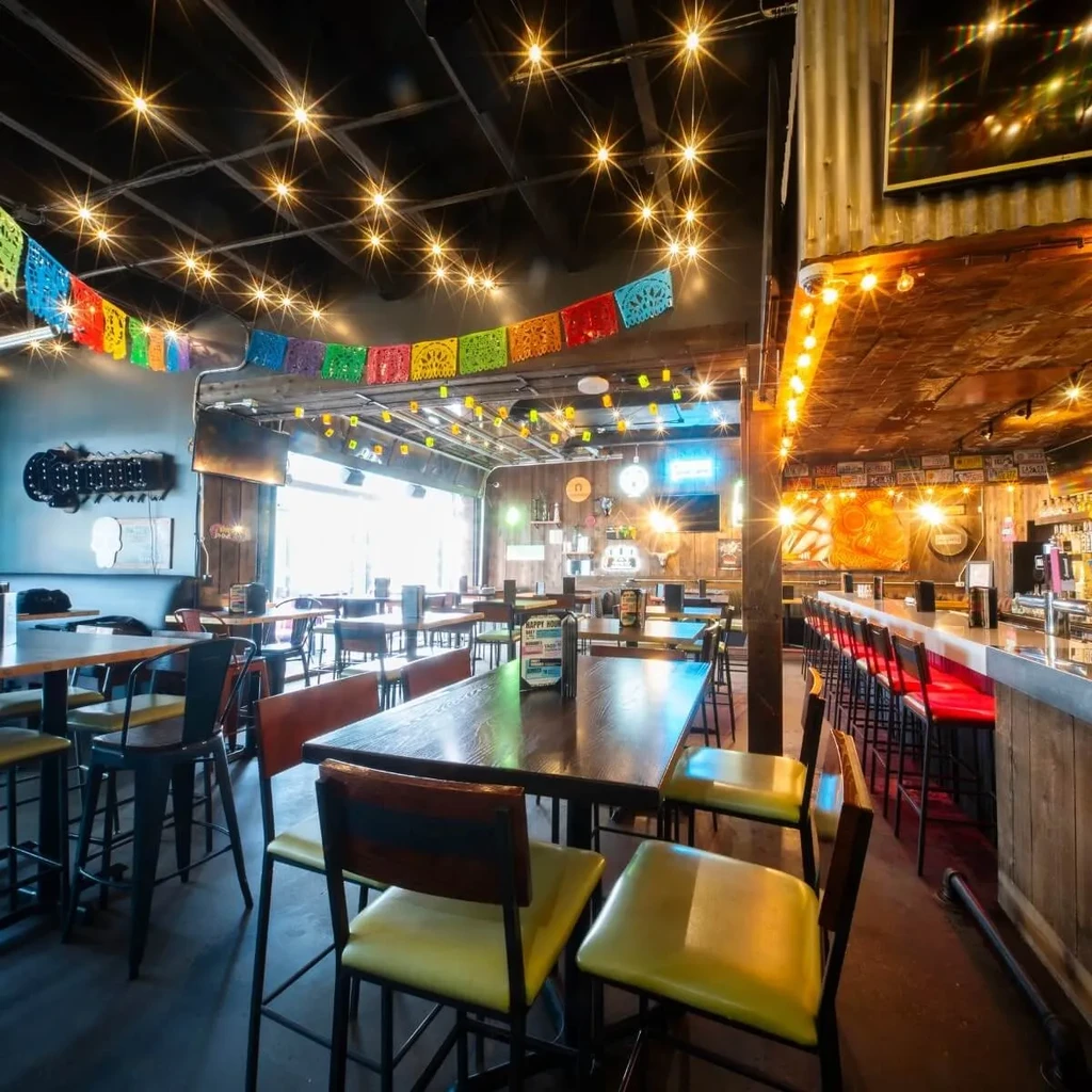 Restaurant interior with industrial-festive decor, warm lights, and colorful banners.