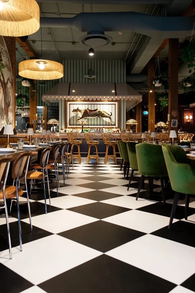 Restaurant interior with checkerboard floor, green velvet chairs, and striped bar.