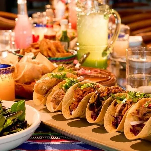 Seven filled tacos, drinks, and side dishes on a colorful table.