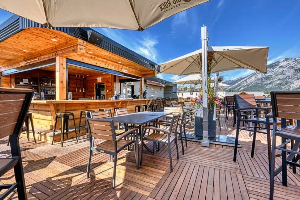 Outdoor restaurant patio bar, wooden deck, umbrellas, snow-capped mountains.