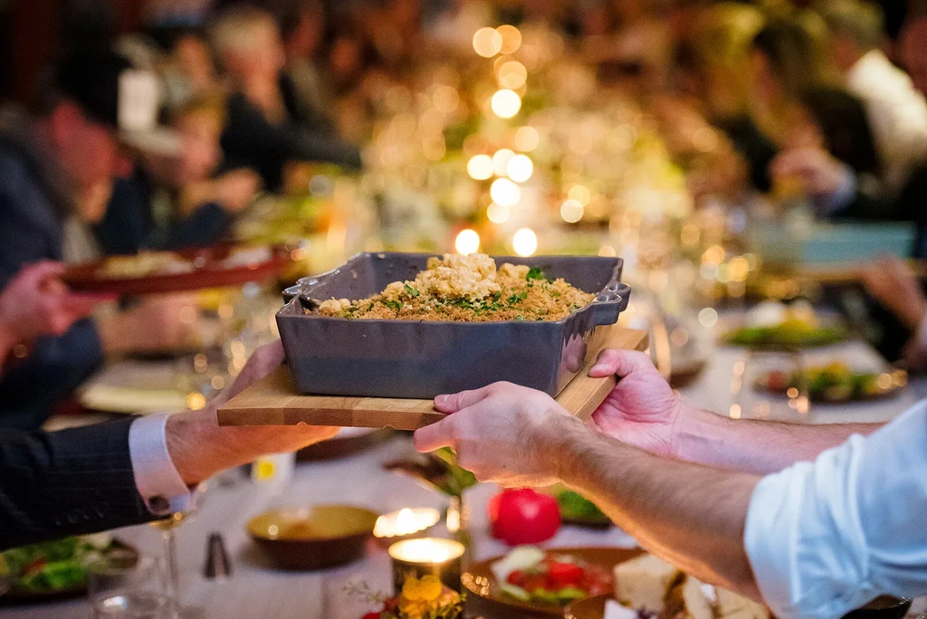 Hands carry a golden-brown baked casserole dish through a warm dining scene.